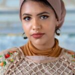 Portrait of a woman in a colorful embroidered outfit, wearing a headscarf and hoop earrings, looking directly at the camera.